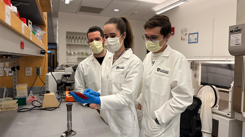 students in white lab coats, wearing masks. One student is warming a sample over a Bunsen burner