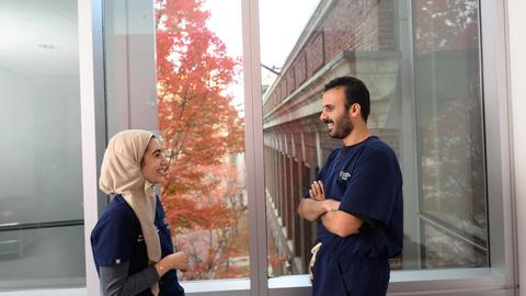 Two dental students in scrubs talk beside a window overlooking colorful autumn foliage.