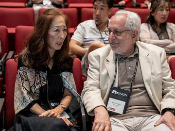 Two attendees talk while seated in an auditorium before a session.