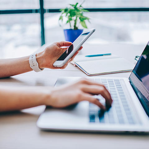 woman typing on a laptop with a cell phone in one hand.