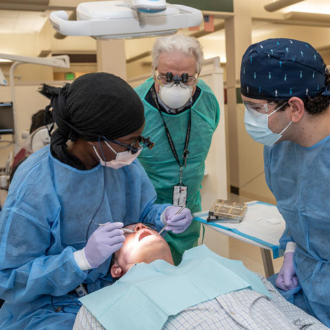 patient in a clinical setting. The students are wearing blue surgical gowns, masks, and gloves, while the instructor supervises closely.