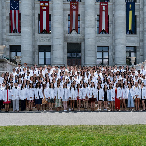 Class of 2028 in their white coats pose on the stairs at the main entrance to HSDM.