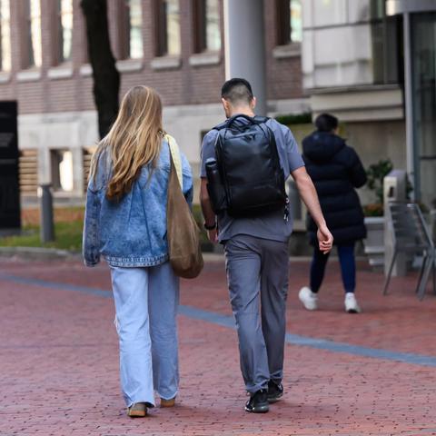 Students walk along a brick pathway on campus in autumn, with red and orange foliage, academic buildings, and a modern glass entrance in the background.