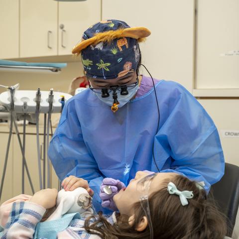 Dental student in PPE and wearing lion ears leans over a child in a dental chairl. Child has her mouth wide open.