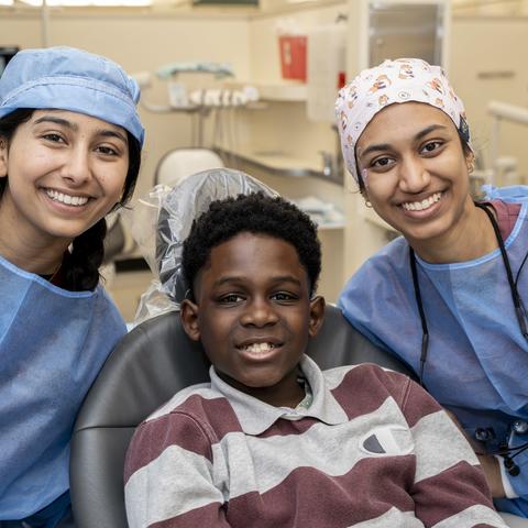 Two dental students in PPE smiling flanking a patient in the dental chair also smiling
