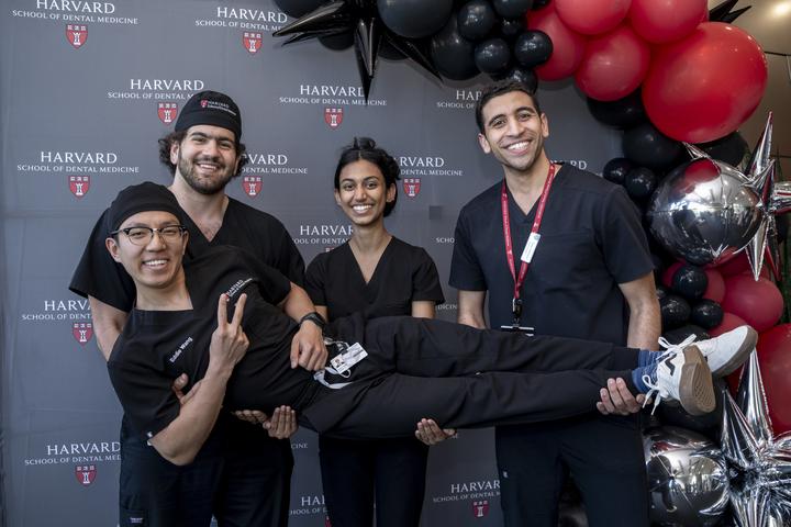 A group of four Harvard dental students in black scrubs pose together in front of a Harvard School of Dental Medicine backdrop, with one student held horizontally while smiling and flashing a peace sign.