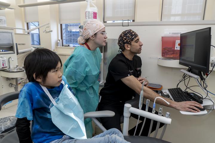 A dental student and a classmate review information on a computer screen while treating a young patient seated in a dental chair during a pediatric dental care event.