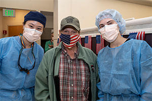 A veteran easing an American flag face mask is flanked by two masked dental strudents.