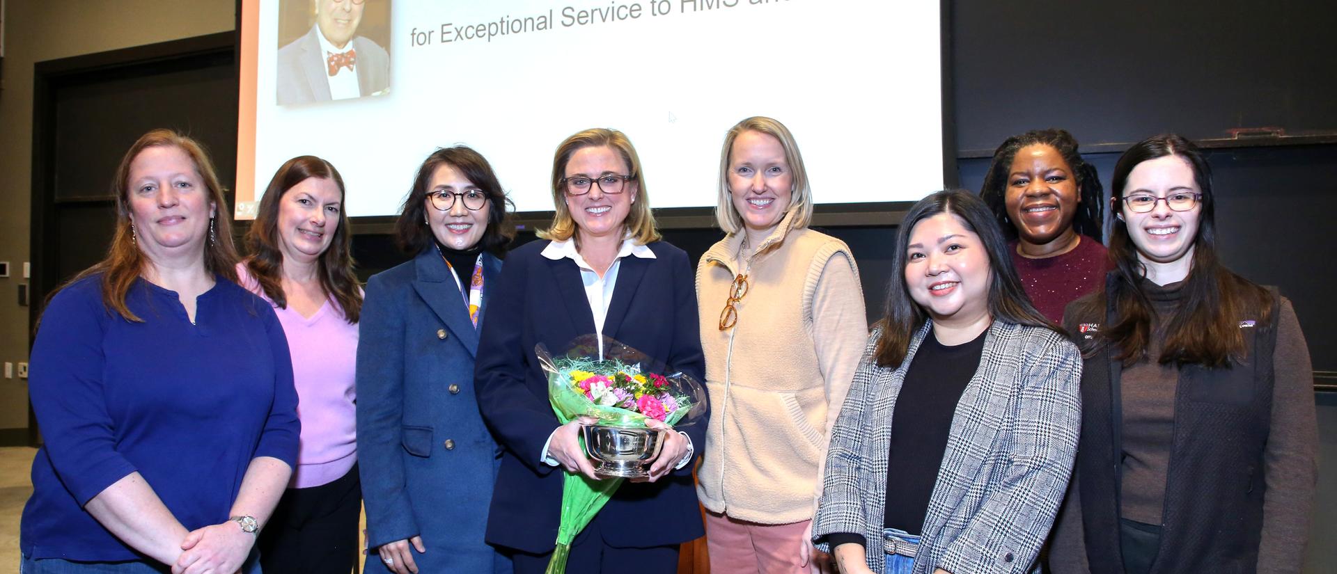 Sara holding a silver bowl and flowers surrounded by her colleagues
