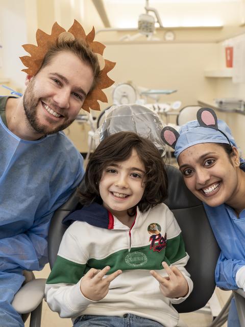 two sudents in PPE flank a child in a dental chair. The students are smiling