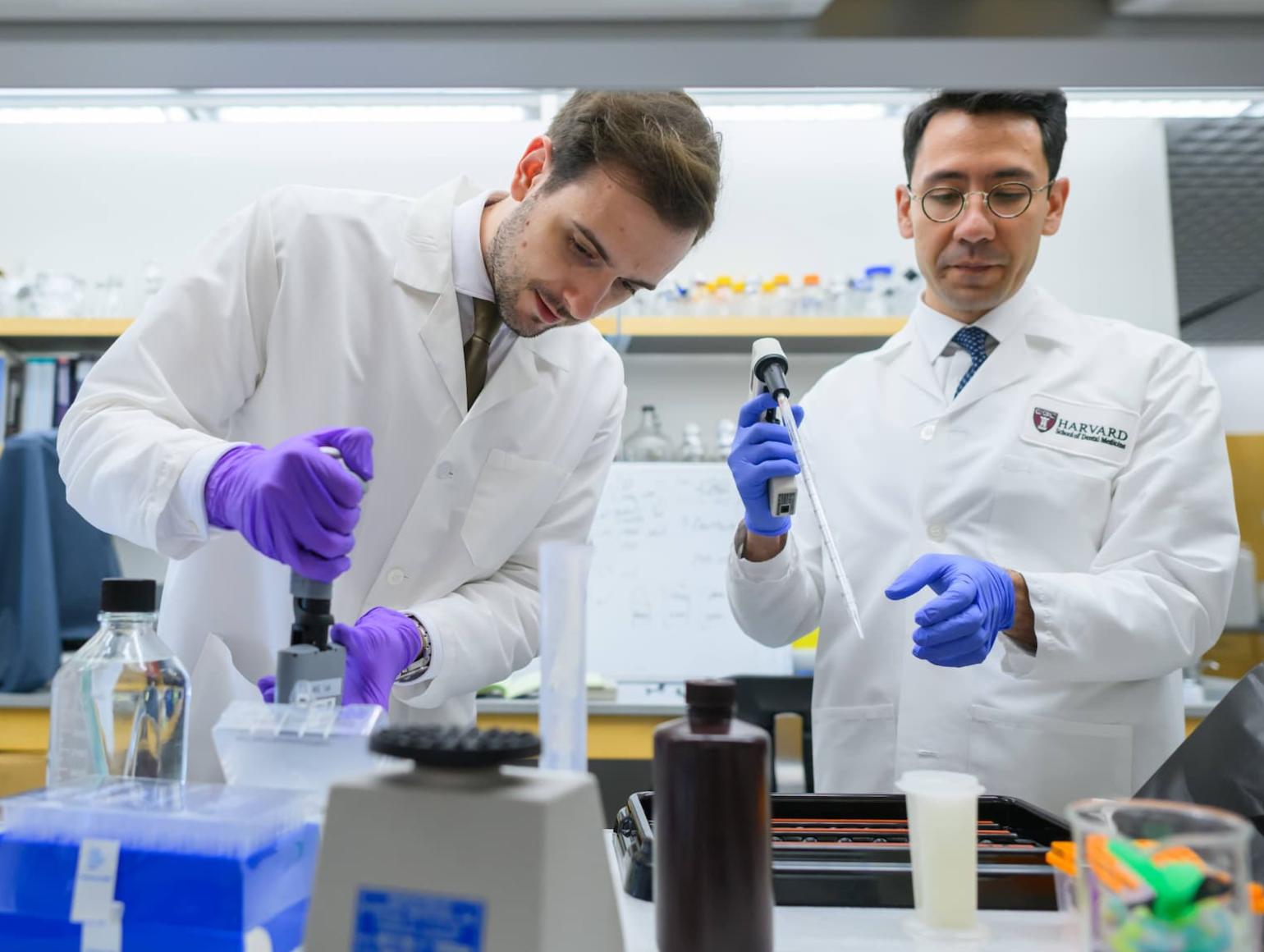 Two researchers in white coats working together both are holding automated pipettes.