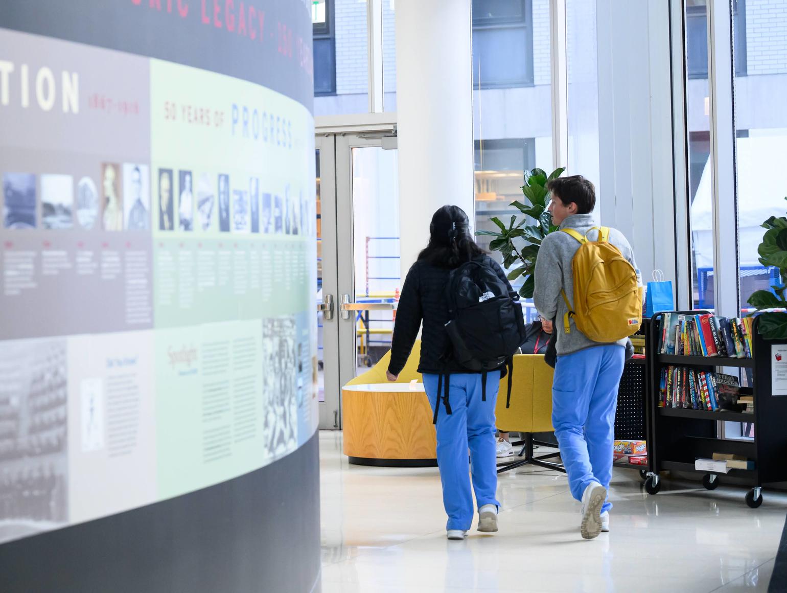 Two students walk through a bright campus lobby near a wall display highlighting the history of Harvard School of Dental Medicine.