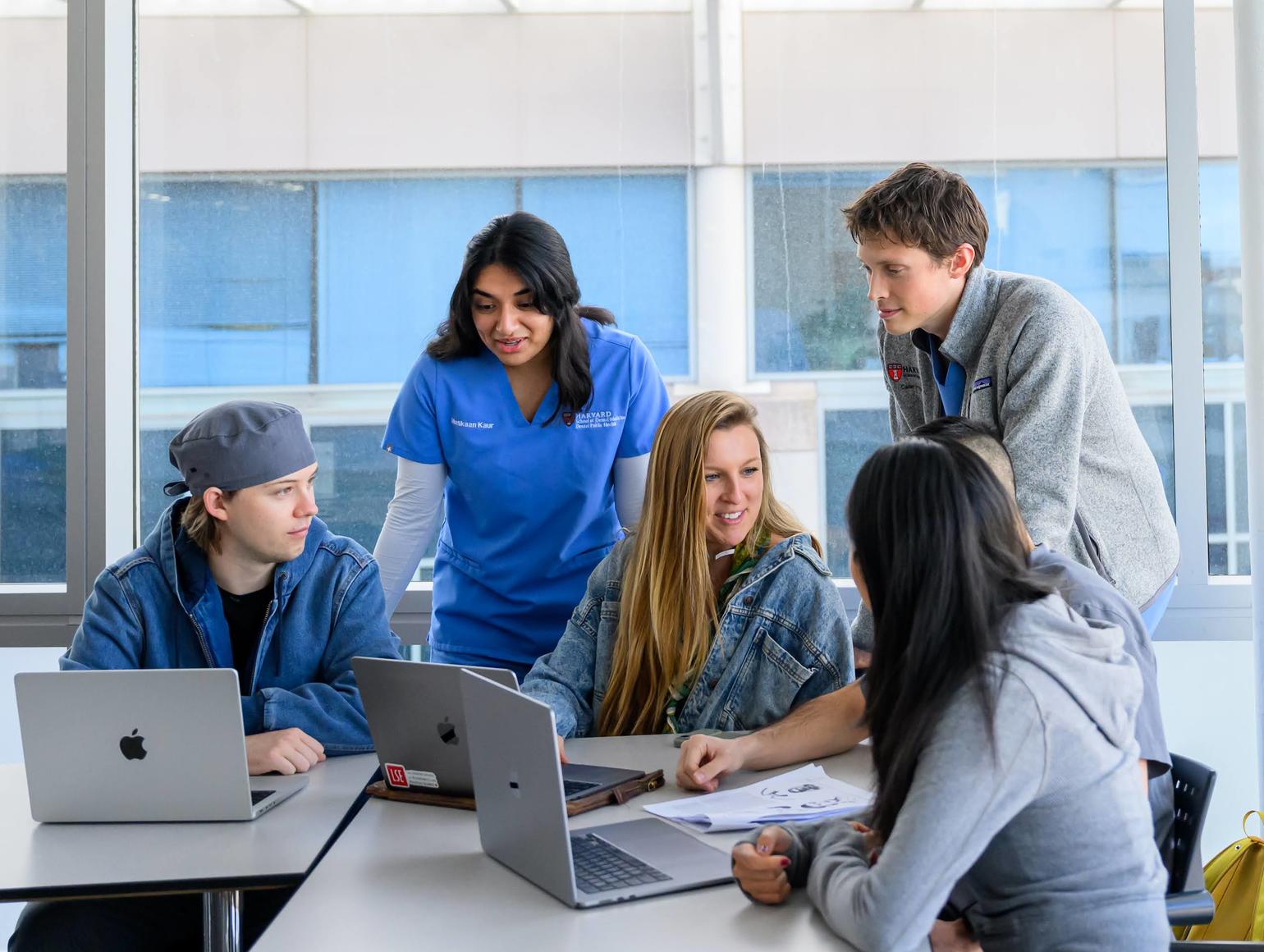 A small group of students gathered around a table in a bright classroom, collaborating and talking while working on laptops.