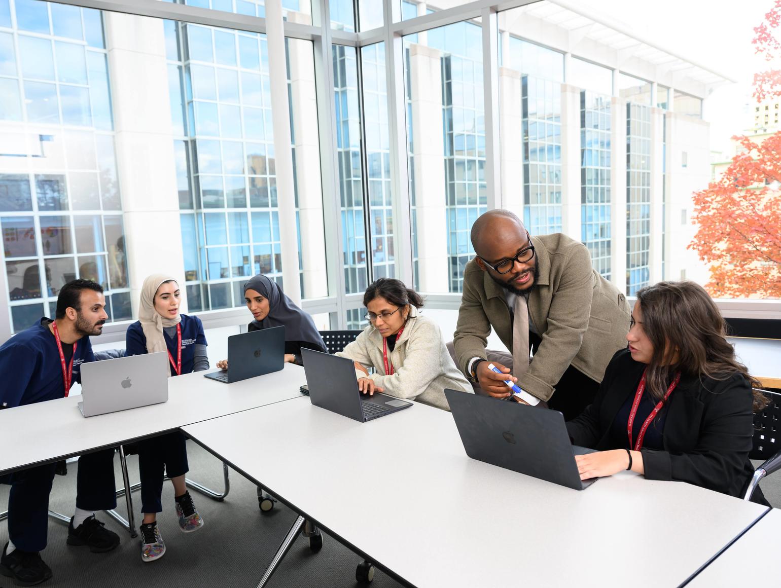 An instructor leans over a table, pointing to a student’s laptop screen while a small group of students work together in a bright classroom.