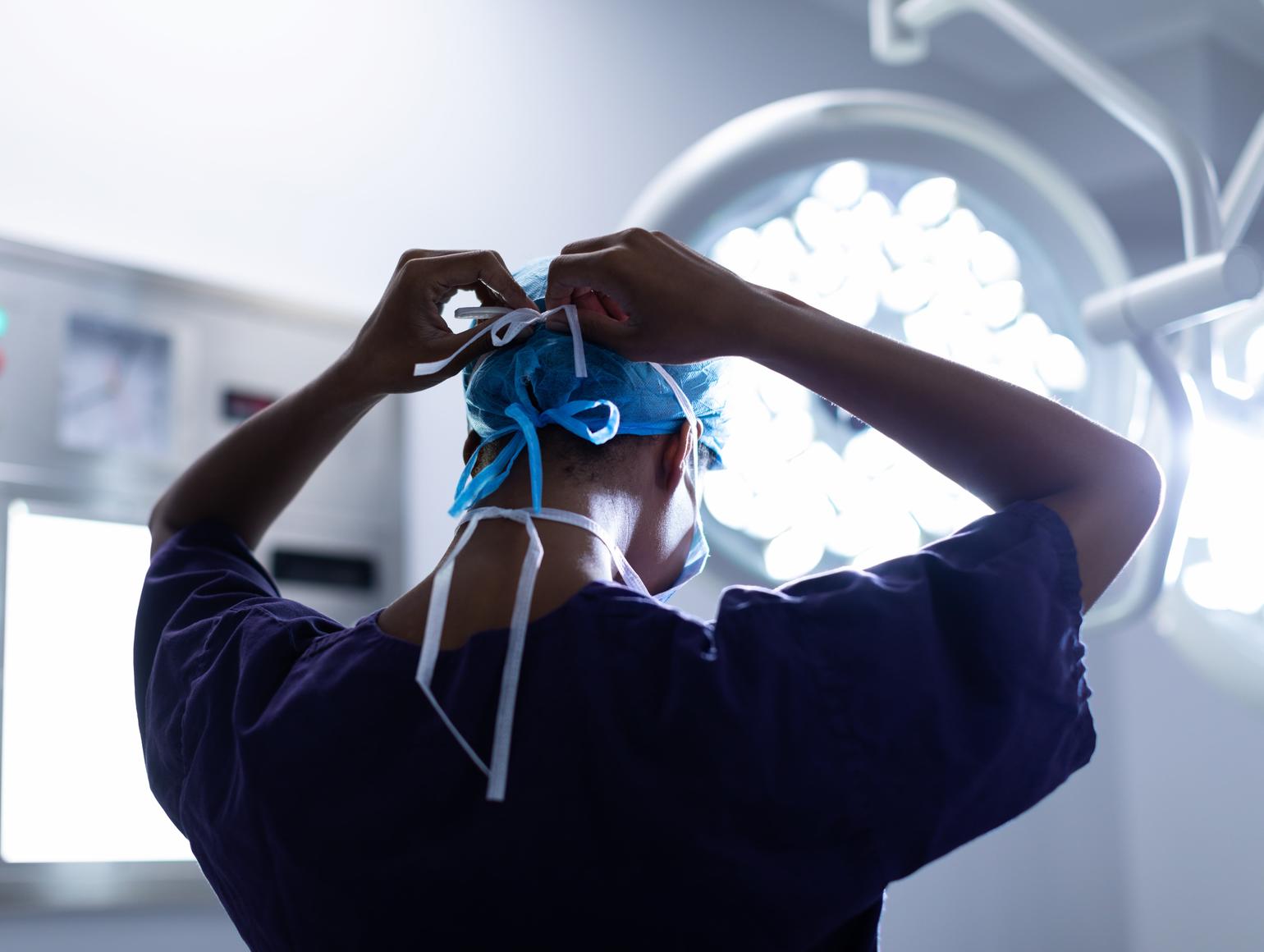 A surgeon standing in front of a large surgical overhead lamp tying on a surgical mask, viewed from behind