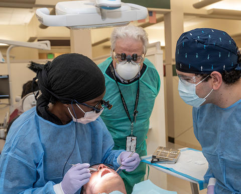 patient in a clinical setting. The students are wearing blue surgical gowns, masks, and gloves, while the instructor supervises closely.