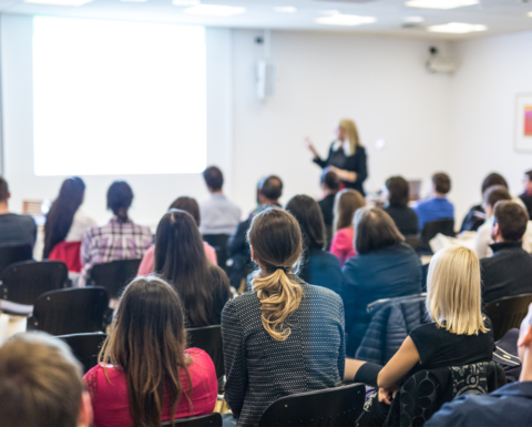 An full auditorium shot from the back of the room with an instructor in the from of the room pointing at a screen 