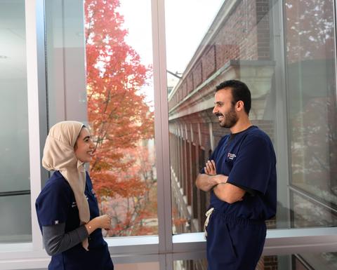 Two dental students in scrubs talk beside a window overlooking colorful autumn foliage.